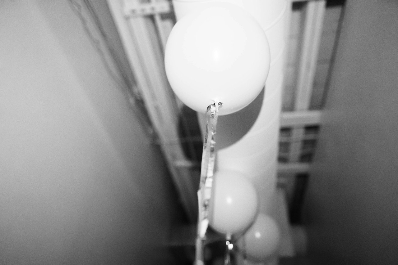 Weiße Luftballons hängen an einer Säule in einem kahlen Innenraum, in Schwarzweiß fotografiert.