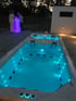 Ein beleuchteter Pool und Whirlpool bei Nacht, mit zwei Personen an einer leuchtenden Bar im Hintergrund.