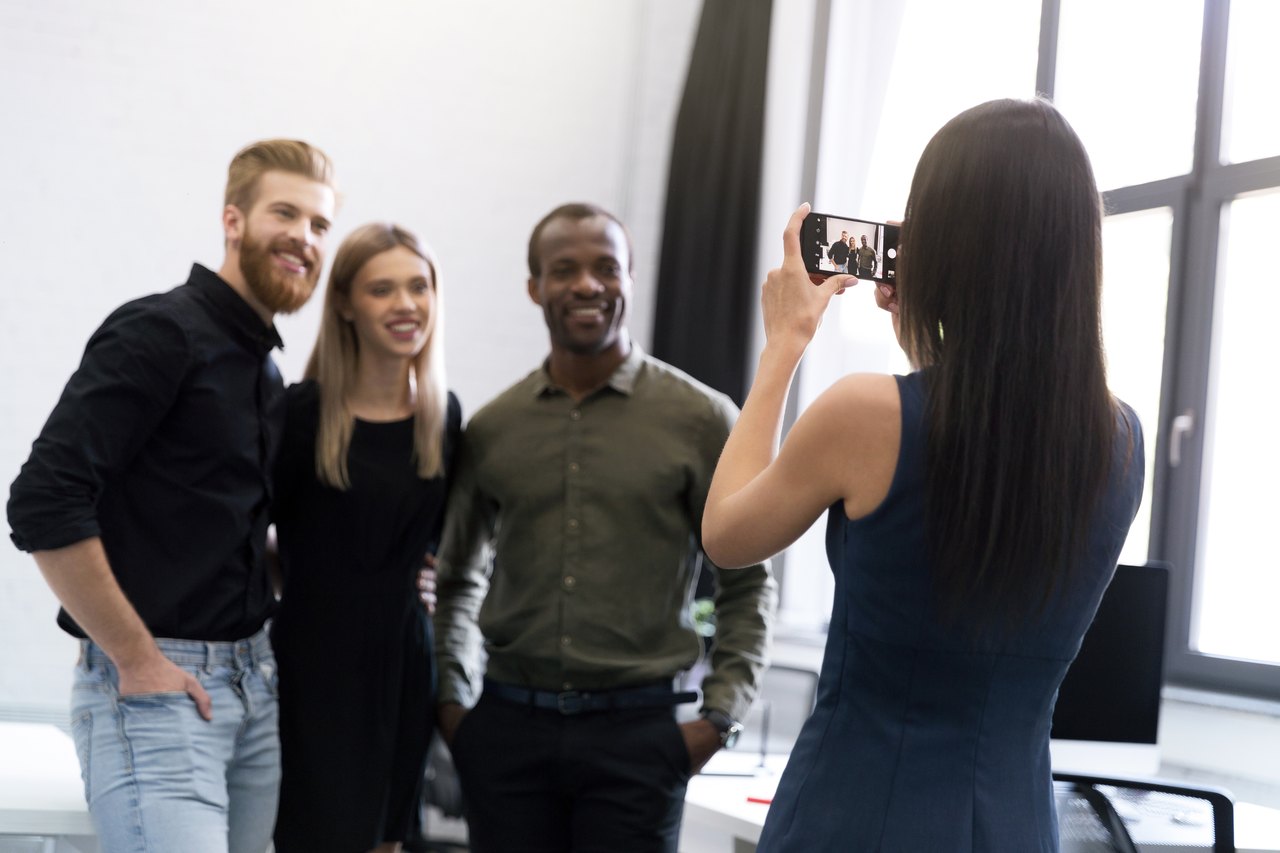 Eine Frau fotografiert drei lächelnde Kollegen im Büro.
