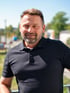 Ein lächelnder Mann mit kurzem braunem Haar und Dreitagebart trägt ein schwarzes Poloshirt vor hellem Außenhintergrund.