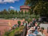 Tennisanlage mit roten Sandplätzen, Zuschauern auf der Terrasse und einem historischen, runden Wasserturm im Hintergrund unter einem sonnigen Himmel.