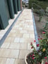 Gepflasterter Gartenweg aus beigen Steinplatten mit dunkler Randeinfassung neben einer Hauswand und Blumenbeeten.