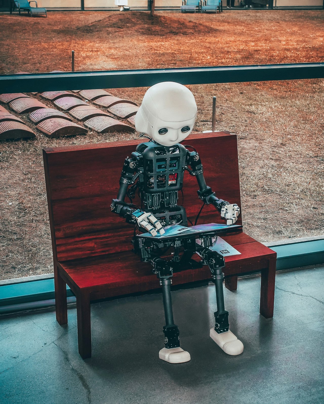 Ein kleiner humanoider Roboter mit weißem Kopf sitzt auf einer Holzbank und bedient ein Tablet.