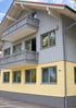 Mehrstöckiges Wohnhaus mit gelber Putzfassade im Erdgeschoss und grauer Holzverkleidung mit Balkonen in den Obergeschossen.