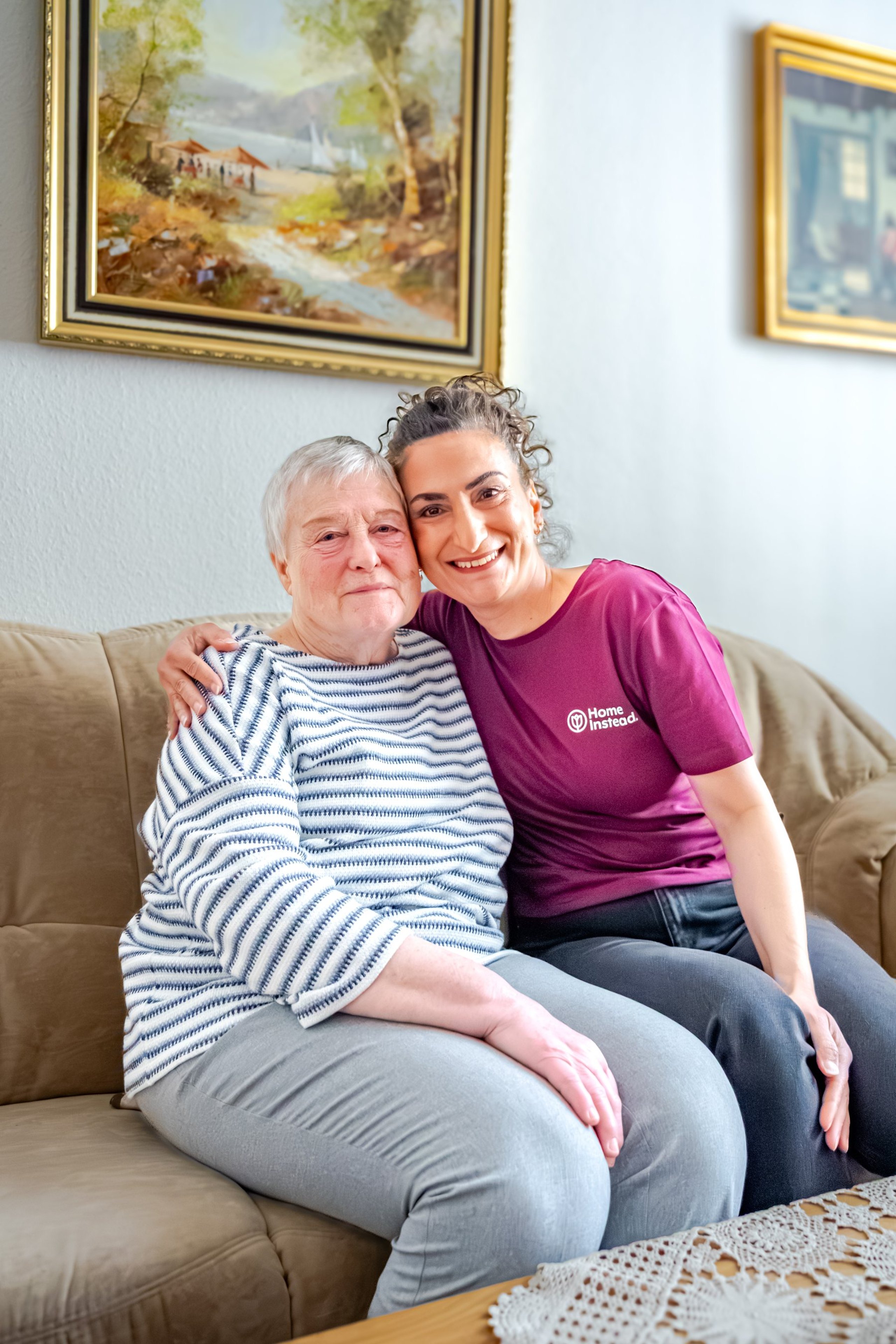 Betreuungskraft von Home Instead mit Seniorin, Arm in Arm auf dem Sofa