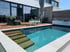 Moderner Gartenpool mit Treppeneinstieg, Holzterrasse, Pflanzkübeln und Sonnenschirm vor einer Glasfront.