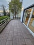 Gepflasterte Terrasse mit grauen Steinplatten, weißem Geländer und Blick auf einen Baum sowie eine Glasfront.