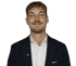Mann mit lockigen, braunen Haaren trägt ein dunkelblaues Sakko mit dezentem Karomuster und weißem Hemd vor weißem Hintergrund.