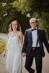 A smiling woman in white overalls holding hands with an older man in a suit and bow tie.
