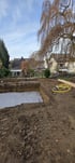 Ausgehobene Baugrube im Garten mit grauer Schutzfolie, gelbem Schlauch und Wohnhäusern im Hintergrund.