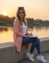 Jana von MommyOnHerTerms  rosa Strickjacke und Jeans sitzt am Flussufer und schreibt in ein Notizbuch bei Sonnenuntergang.