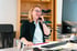 Frau mit Brille am Schreibtisch, lachend, telefoniert, vor einem Regal.