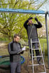 Zwei Männer prüfen eine Schaukel auf einem Spielplatz, einer auf einer Leiter montiert die Kette, der andere notiert.