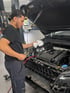 Ein Mechaniker in dunkler Kleidung wischt mit einem weißen Tuch am offenen Motorraum eines Autos.