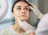 Hände in weißen Handschuhen untersuchen das Gesicht und die Kinnpartie einer jungen Frau bei einer dermatologischen Beratung.