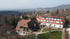 Großes Hotelgebäude mit rotem Dach und Solarpaneelen, umgeben von Bäumen und einem grünen Rasen, mit Hügeln im Hintergrund.
