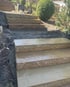 Blockstufen aus beigem Naturstein bilden eine Treppe im Gartenhang neben einem Buchsbaum und frischer Erde.