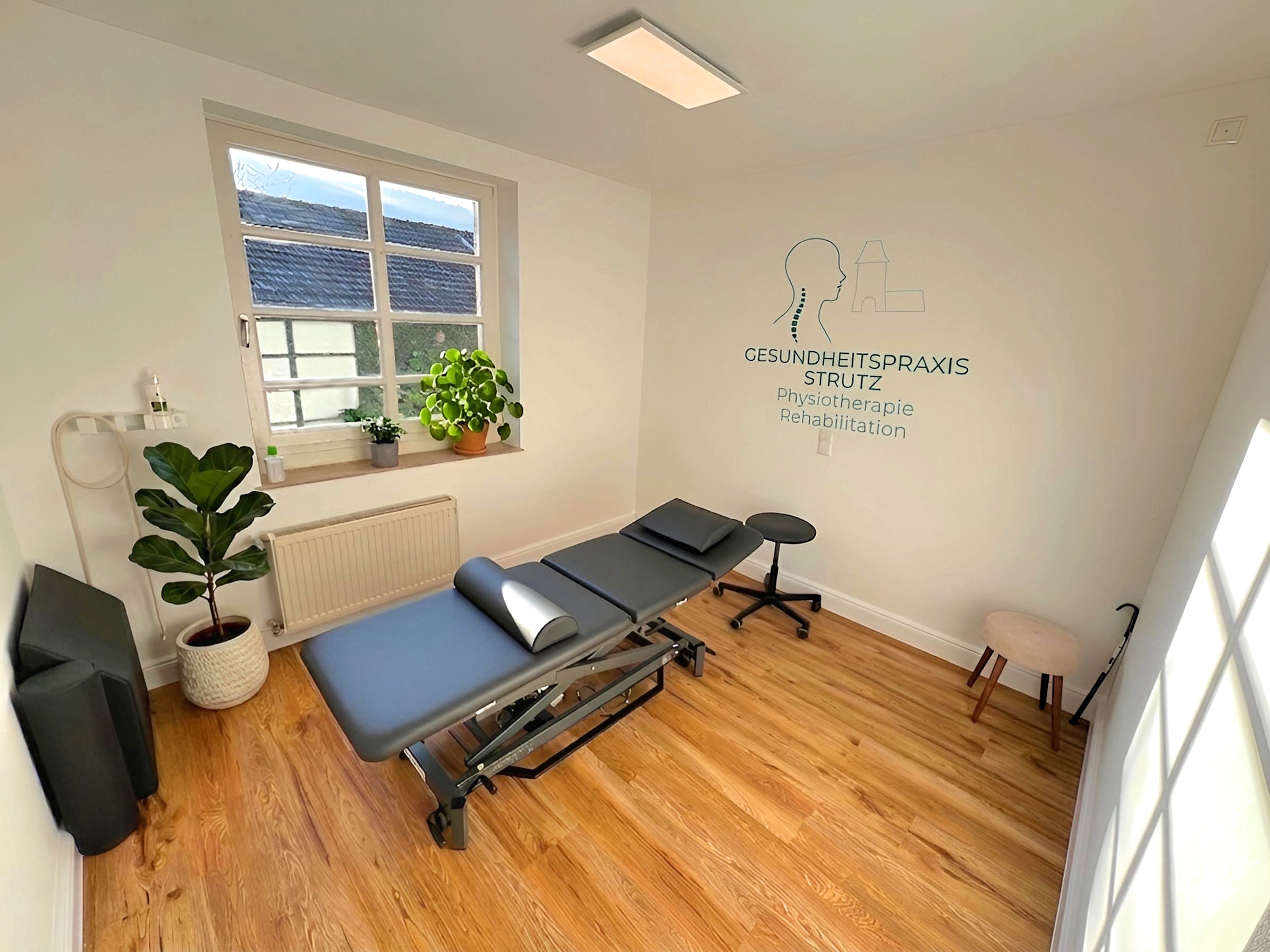 Behandlungsraum mit Liege, Pflanzen und Schriftzug Gesundheitspraxis Strutz an der Wand