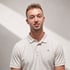 Junger Mann in beigem Poloshirt mit "CK"-Logo, lächelnd vor weißer Wand mit Lichtstreifen.