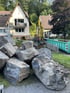 Gelber Bagger arbeitet an einer Baustelle mit großen Steinblöcken vor einem Wohnhaus mit Wendeltreppe und grünem Container.