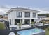 Modernes weißes Einfamilienhaus mit Terrasse, Gartenpool, Whirlpool und Hängematte unter bewölktem Himmel.