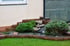 Kleiner Garten-Teich mit Wasserfall, umgeben von grünen Pflanzen, Steinen und Ziegeln, vor heller Hauswand.