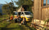 A family relaxes outside next to a gold and white campervan at the campsite with its roof tent open; a woman is looking out.