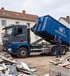 Ein Bausantec LKW entlädt einen blauen Schuttcontainer auf einer Baustelle neben einem Haufen Bauschutt.