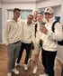 Fünf lächelnde Männer in cremefarbenen Troyer-Pullovern machen ein gemeinsames Spiegelselfie in einem modernem Innenraum.