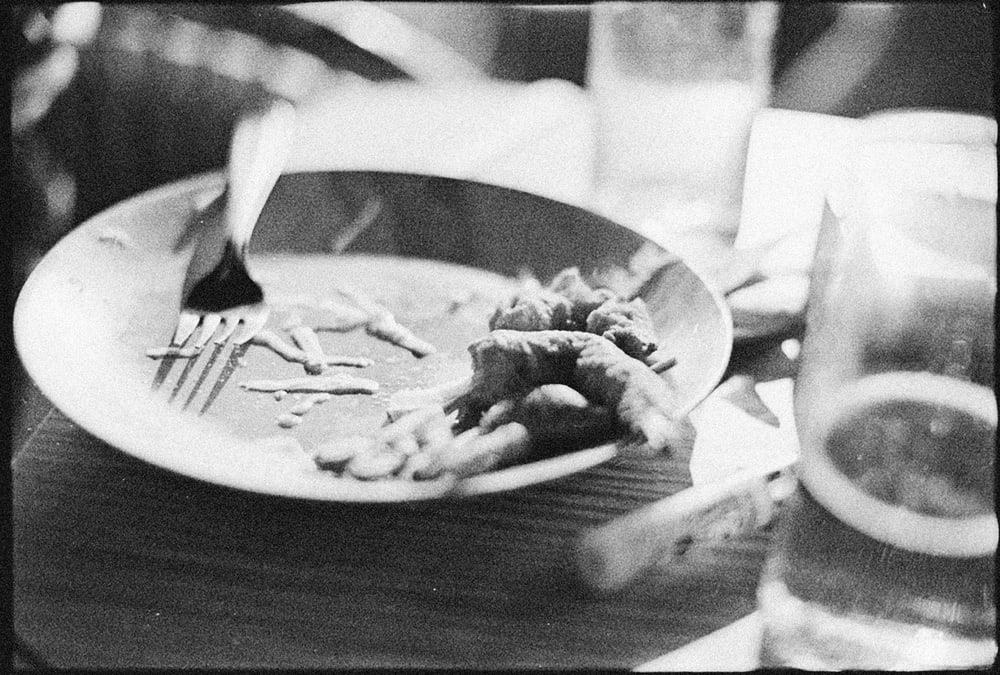 Ein halb leer gegessener Teller mit einer Gabel darin, daneben ein Glas, körnig in Schwarzweiß fotografiert.