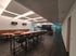 Modern meeting room with long wooden table, bar stools, orange bench, plants and large screen.