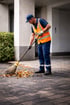Ein Mann in blauer Uniform und orangefarbener Warnweste fegt mit einem Besen Herbstlaub von einem gepflasterten Weg.