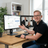 Mann zeigt auf einen Monitor mit Diagrammen und Tabellen, daneben ein zweiter Monitor mit einer Videokonferenz.