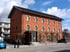 Rotes Backsteingebäude mit mehreren Fenstern und grünen Fensterläden, davor zwei Personen und geparkte Autos.
