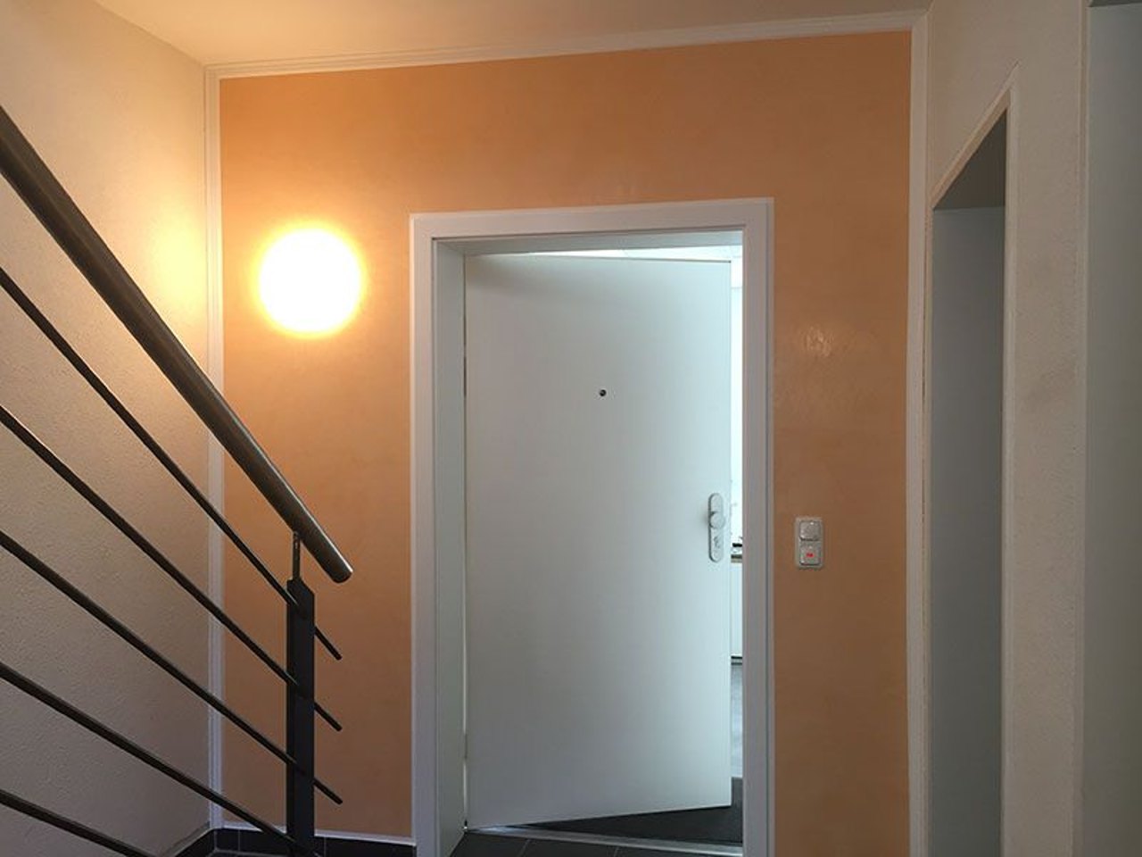 Ein heller Hausflur mit einer apricotfarbenen Wand, einer leuchtenden Wandlampe, Treppengeländer und offener weißer Tür.