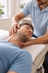 Physiotherapeut behandelt den Nacken eines Mannes, der mit geschlossenen Augen auf einer Couch liegt.