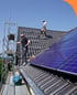 Handwerker installieren Solarmodule auf einem Ziegeldach, mit Gerüst und blauer Plane im Vordergrund.