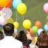 Menschen lassen bunte Luftballons (gelb, orange, blau, rosa, weiß, grün) in den Himmel steigen.