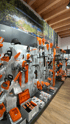Ein Ausstellungsraum mit orange-weißem STIHL-Gartengerät, Zubehör und Batterien auf Regalen unter einem Naturwandbild.