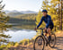 A woman in a blue jersey and helmet rides a bicycle on a dirt path beside a calm lake with mountains in the background.
