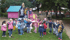 Kinder und zwei Erwachsene spielen im Freien auf einem Spielplatz mit rosa und blauen Geräten.