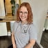 Junge Frau mit rotblonden Haaren, Brille und grauem Polo-Shirt mit "Fitness for Females"-Logo lächelt.
