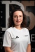 Lächelnde Frau mit Brille im weißen Adidas Sportshirt mit dem Logo von THERAPIER vor dunklem Hintergrund und Pier5 
