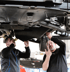 Zwei Mechaniker mit dunklen Jacken reparieren ein Auto von unten in einer Werkstatt.