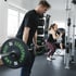 Mann hebt Hantelstange mit Gewichten, im Hintergrund trainiert eine Frau mit Medizinball.