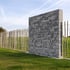 Hohe graue Steinmauer in Kombination mit einem Holzzaun aus vertikalen Staken auf einer grünen Wiese unter blauem Himmel.