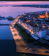 Luftaufnahme einer beleuchteten Küstenstadt bei Dämmerung, mit einem Hafen, Gebäuden und Lichtern entlang der Promenade in Biograd na Moru.