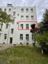 Weißes Mehrfamilienhaus mit Garten, wobei zwei Fenster im ersten Stock mit einem roten Rahmen markiert sind.