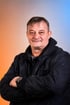 Mann mit dunkler Jacke und verschränkten Armen vor einem orange-blauen Hintergrund.