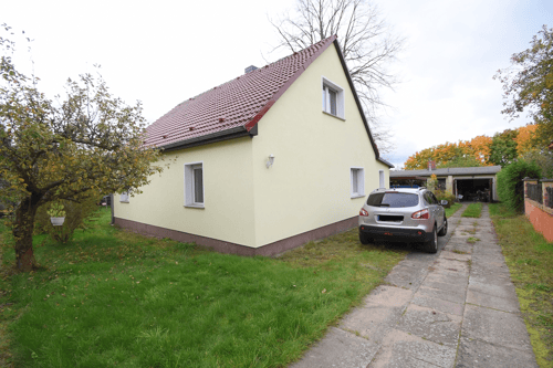 Ein hellgelbes Haus mit rotem Dach und einem silbernen SUV auf einer gepflasterten Auffahrt neben Rasenflächen.